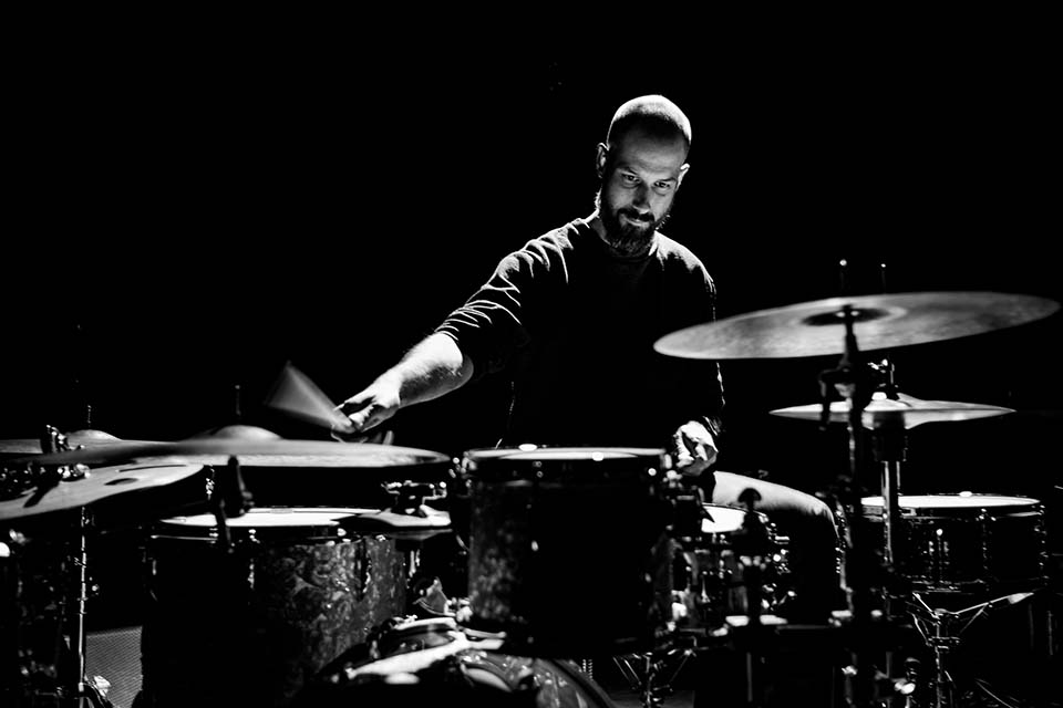A black and white image of a man with a beard, playing the cymbals and performing on a drum kit.